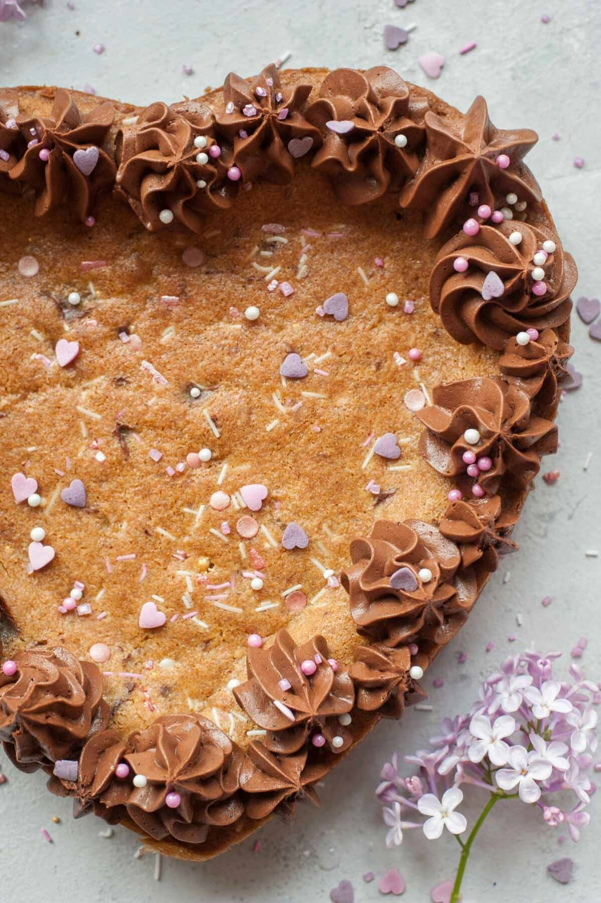 Chocolate chip cookie cake with chocolate frosting on a grey background. Sprinkles and flowers scattered around.