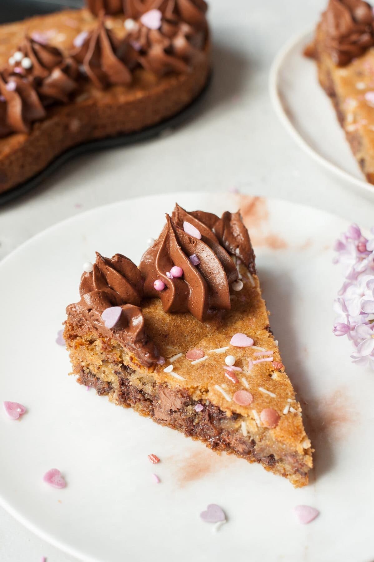 A piece of chocolate chip cookie cake with chocolate frosting on a white plate. Sprinkles and flowers scattered around.