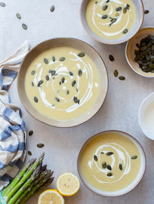 Three bowls with cream of asparagus soup topped with goat cheese and pepitas.