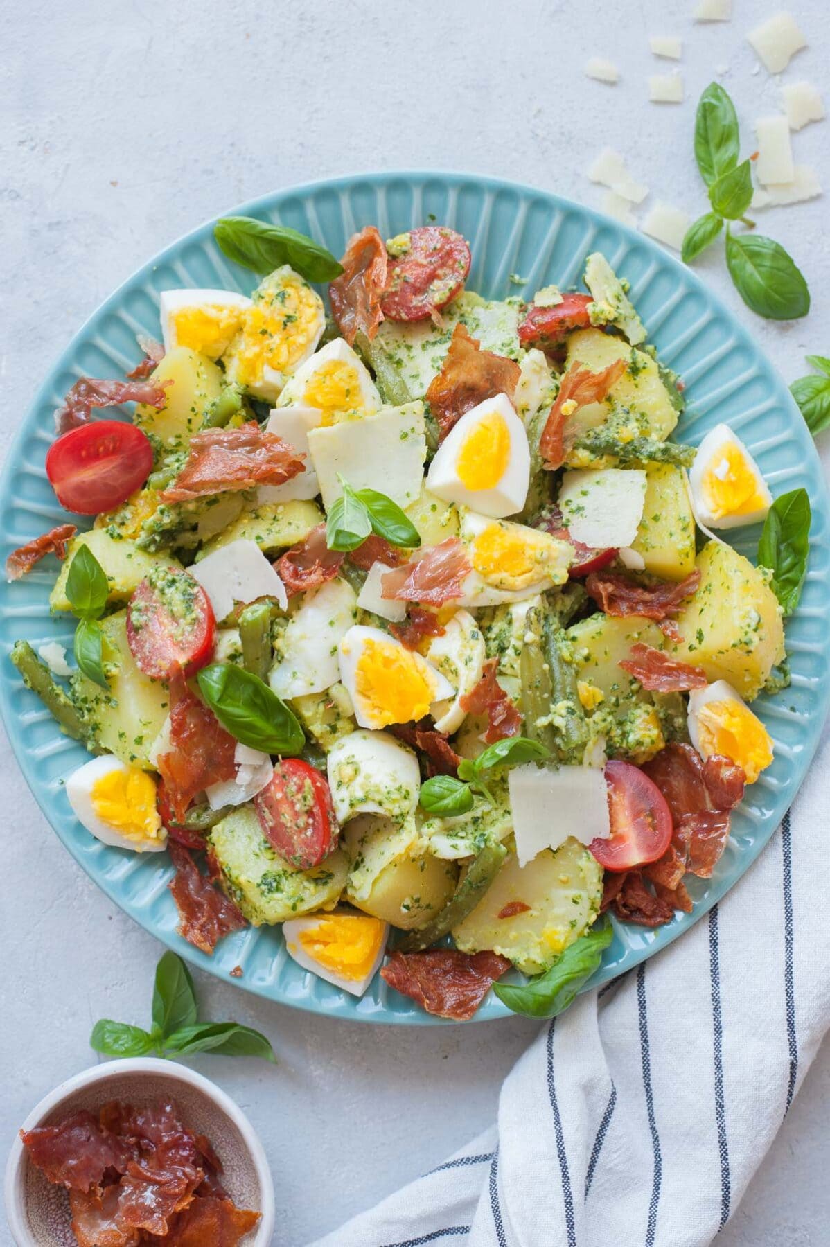 Sałatka ziemniaczana z pesto, jajkami i fasolką szparagową, pomidorkami cherry i szynką prosciutto na niebieskim talerzu.