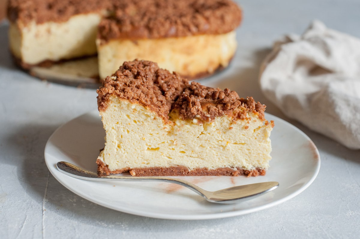 A piece of sernik on a white plate with a spoon. The whole cheesecake in the background.