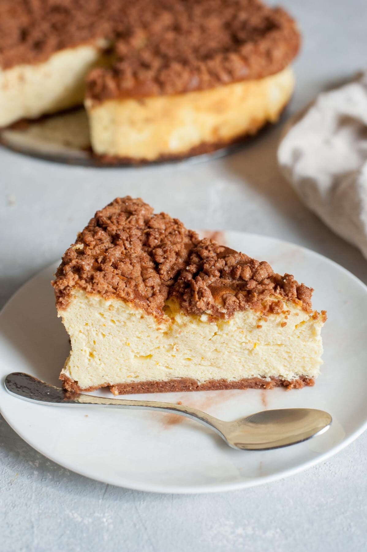 A piece of sernik on a white plate with a spoon. The whole cheesecake in the background.