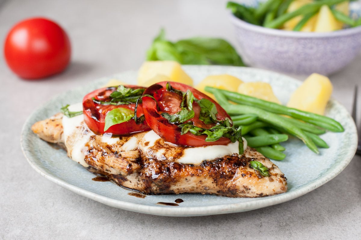 Caprese chicken with mozzarella, tomatoes and basil on a green plate with a side of green beans and potatoes.