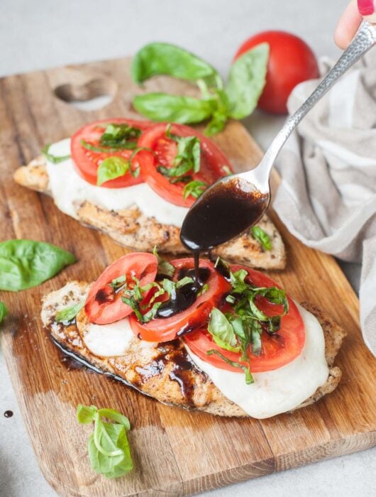 Caprese chicken on a wooden board is being drizzled with balsamic glaze.