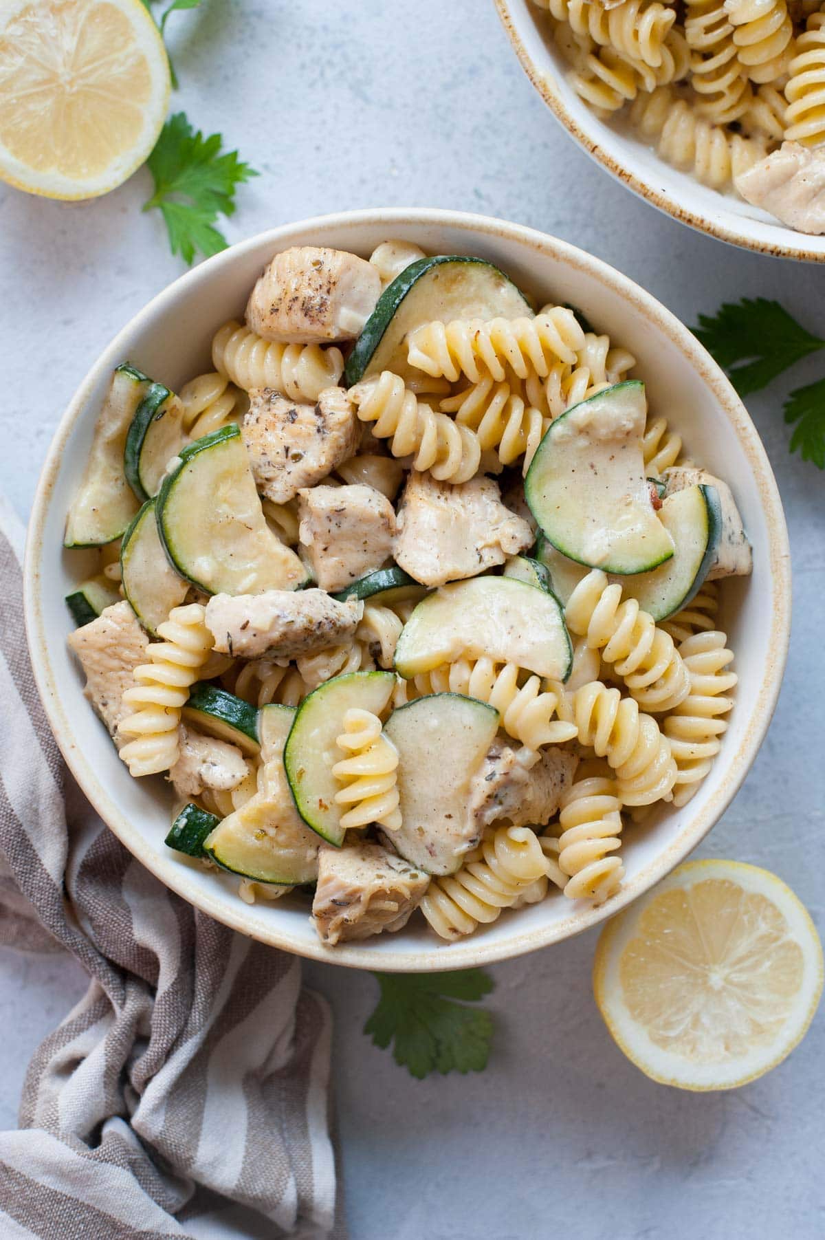 Chicken zucchini pasta in a white bowl. Lemons, parsley, and kitchen cloth in the background.