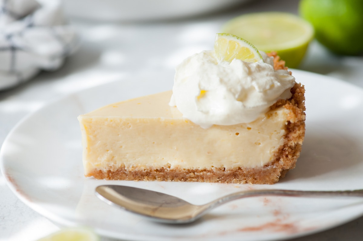 A piece of key lime pie on a white plate. A pie dish and limes in the background.