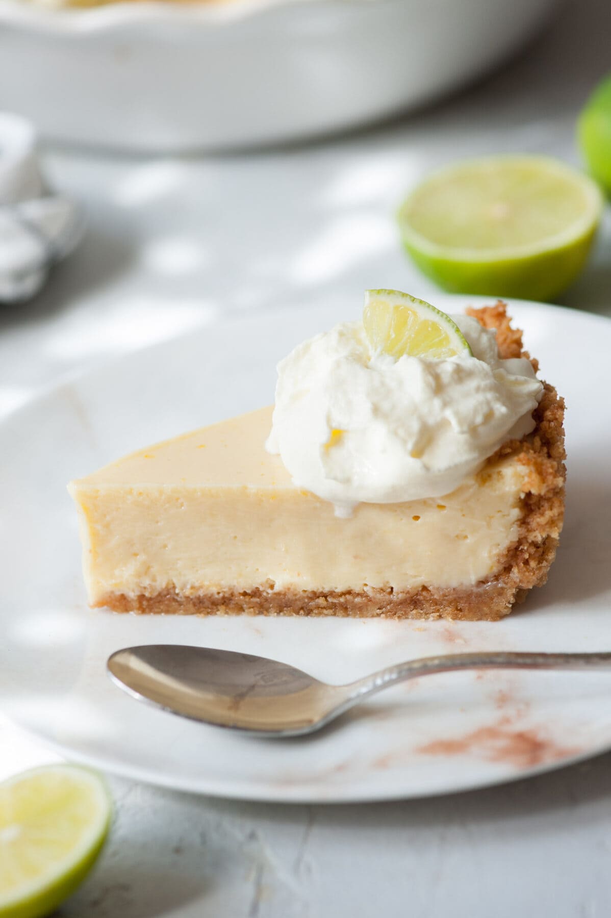 A piece of key lime pie on a white plate. A pie dish and limes in the background.