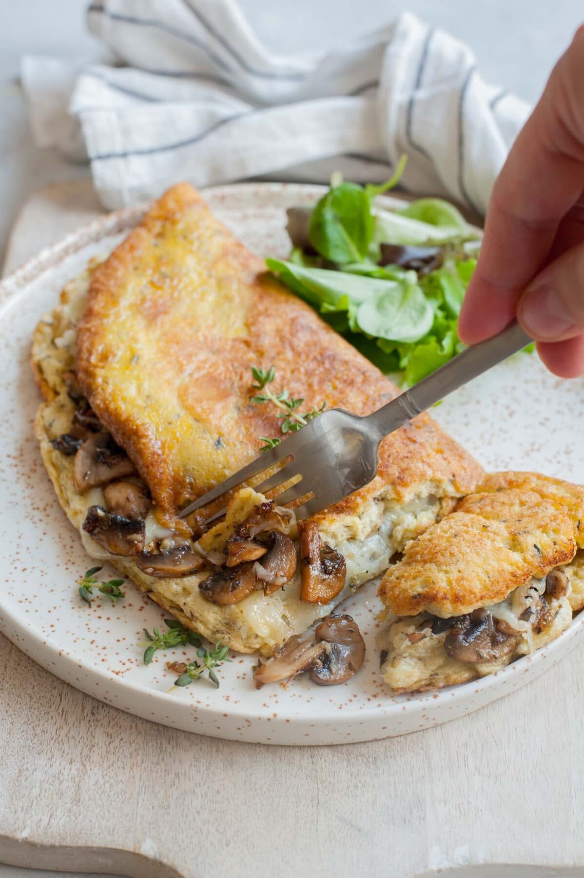 Fluffy omelet filled with mushrooms and cheese on a white plate is being cut with a fork.