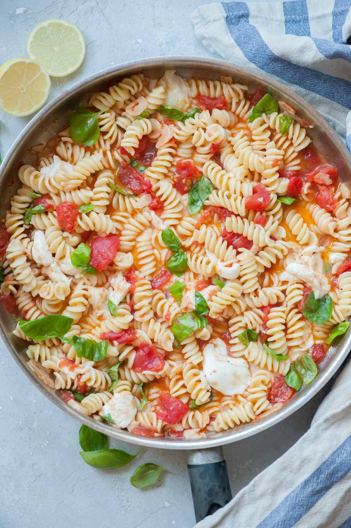 Pasta Caprese in a frying pan. Kitchen cloth, lemons, and basil leaves in the background.