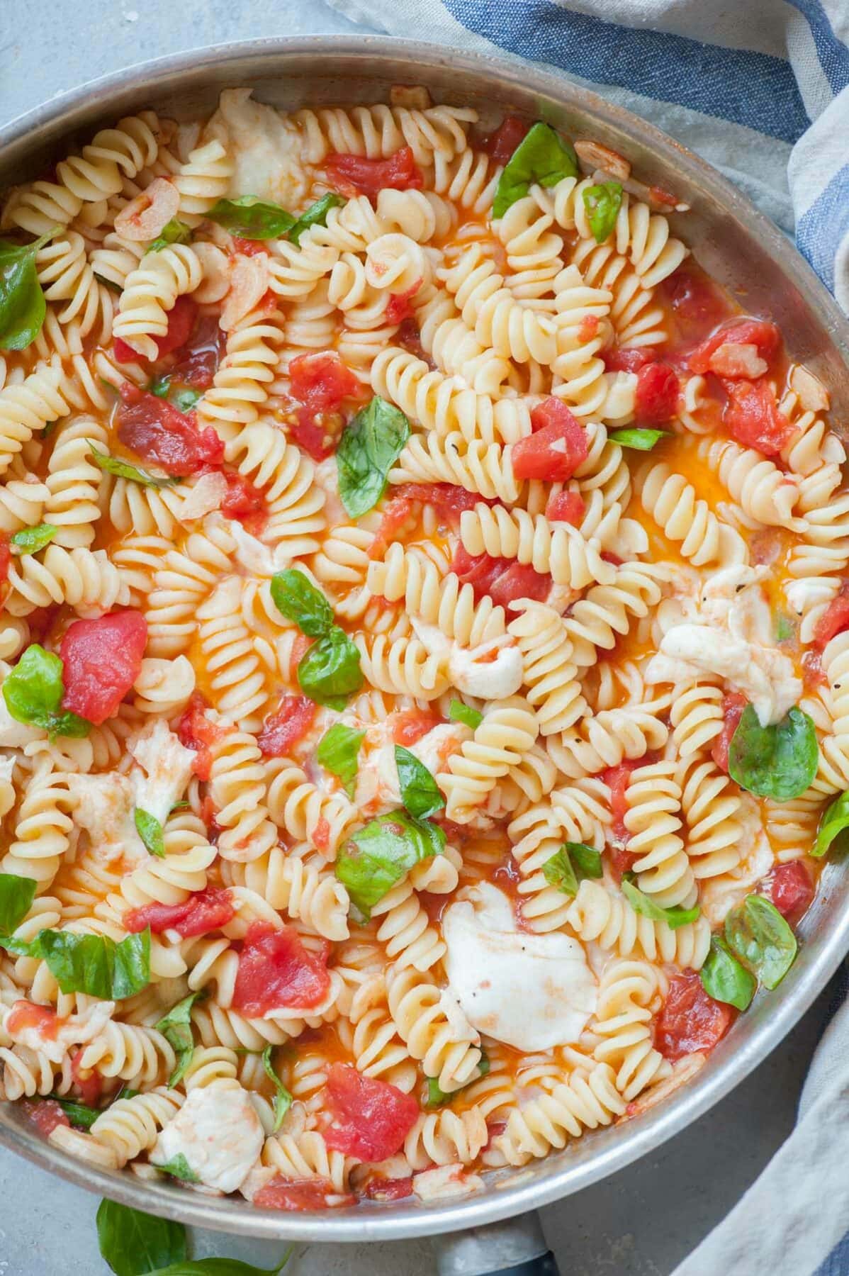 Pasta Caprese in a frying pan. Kitchen cloth and basil in the background.