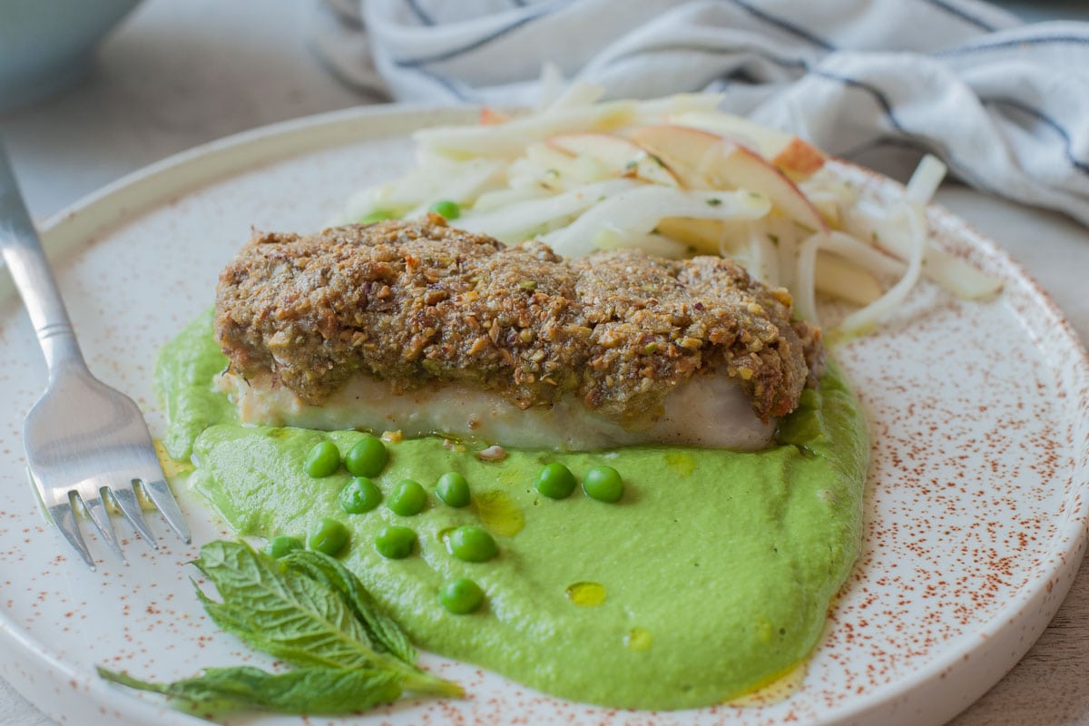 A horizontal picture of pistachio crusted fish with pea puree and kohlrabi slaw on a white plate.