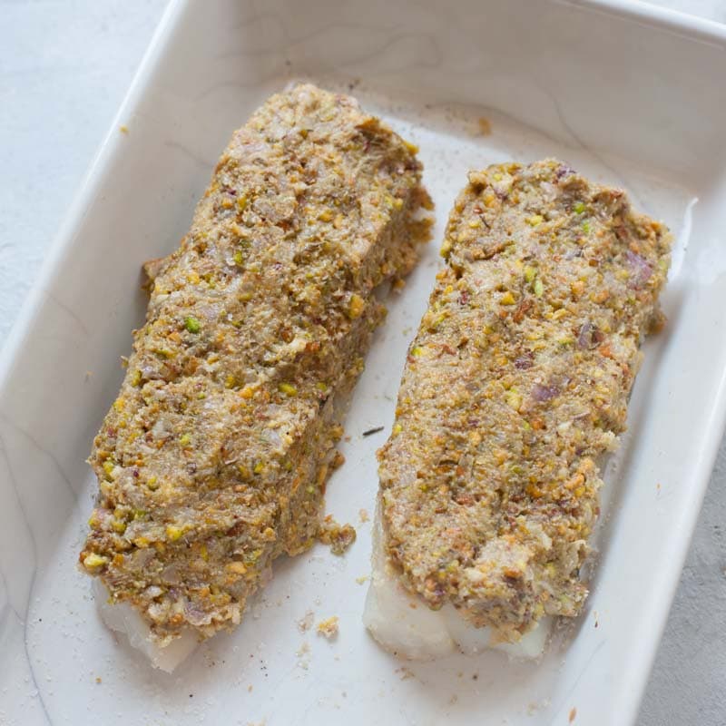 Cod fillets in a white baking dish ready to be baked topped with pistachio mixture. 