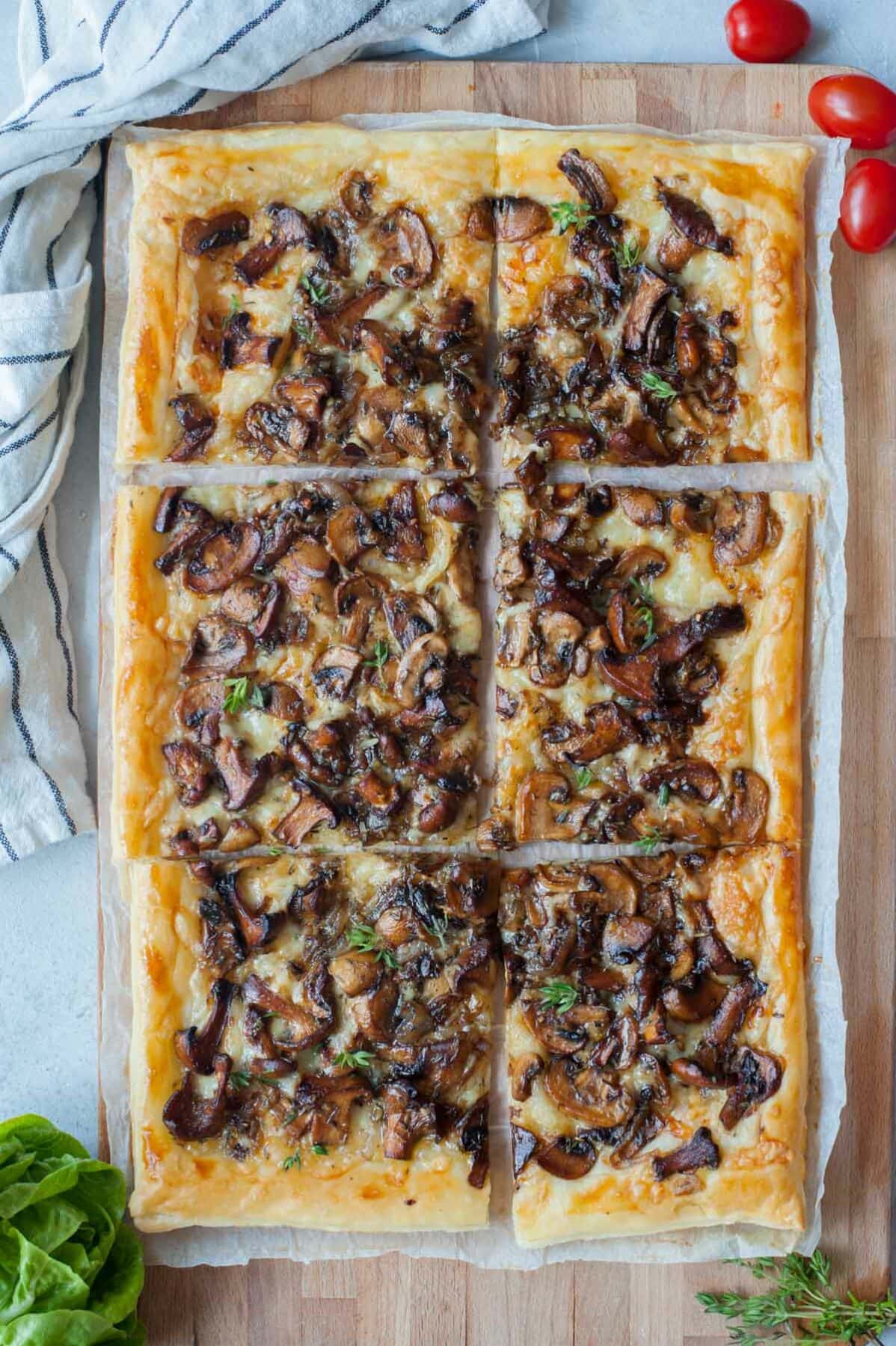 Puff pastry mushroom tart on a wooden board. Tomatoes, thyme, salad, and a kitchen cloth in the background.