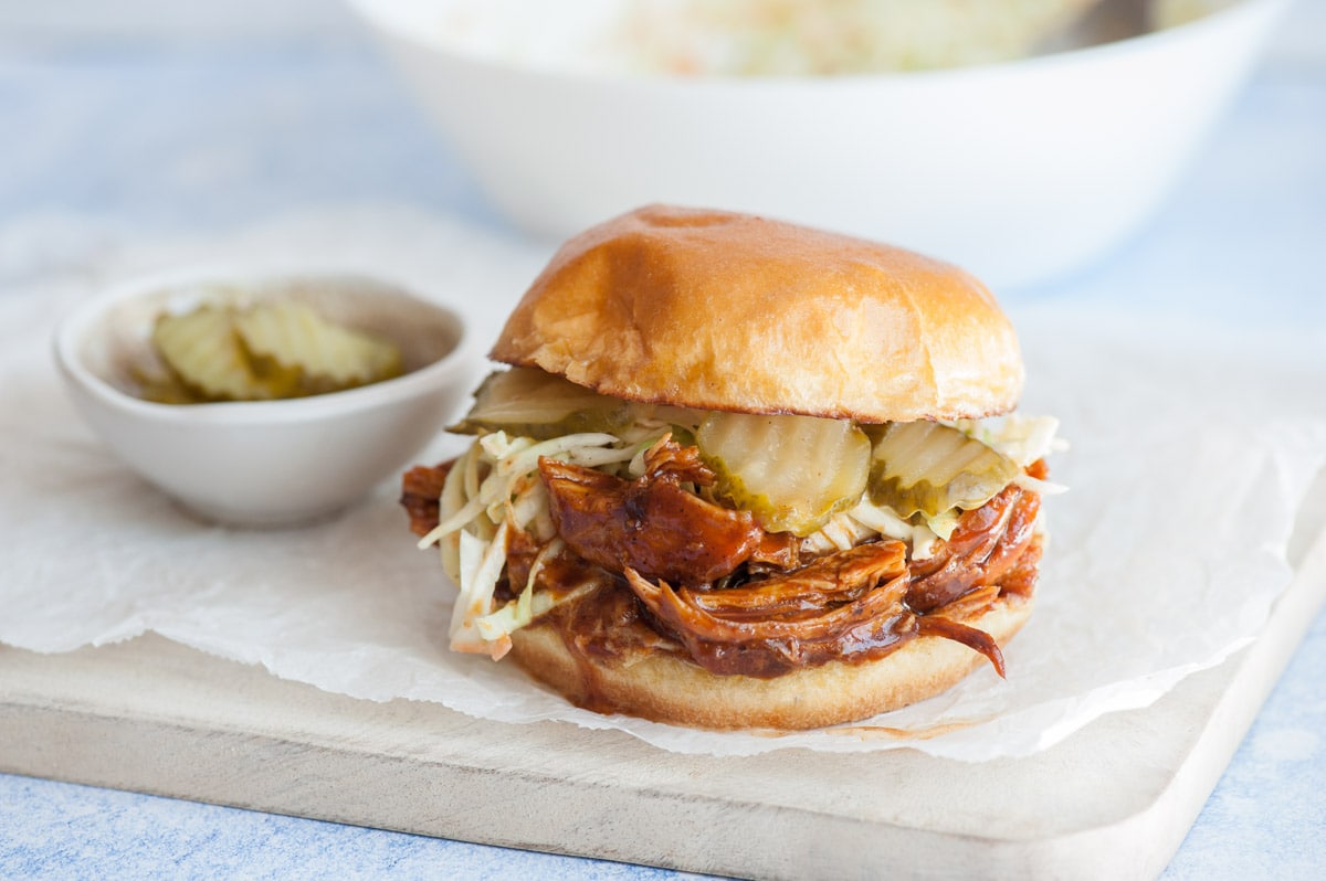 BBQ Pulled Chicken Sandwich on a piece of parchment paper on a white wooden board. Pickles in the background.