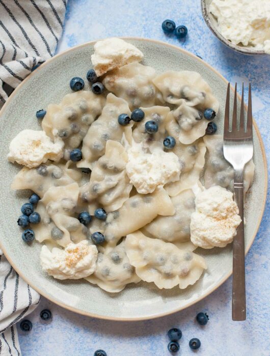 Blueberry pierogi served with fresh blueberries and whipped cream on a blue plate.