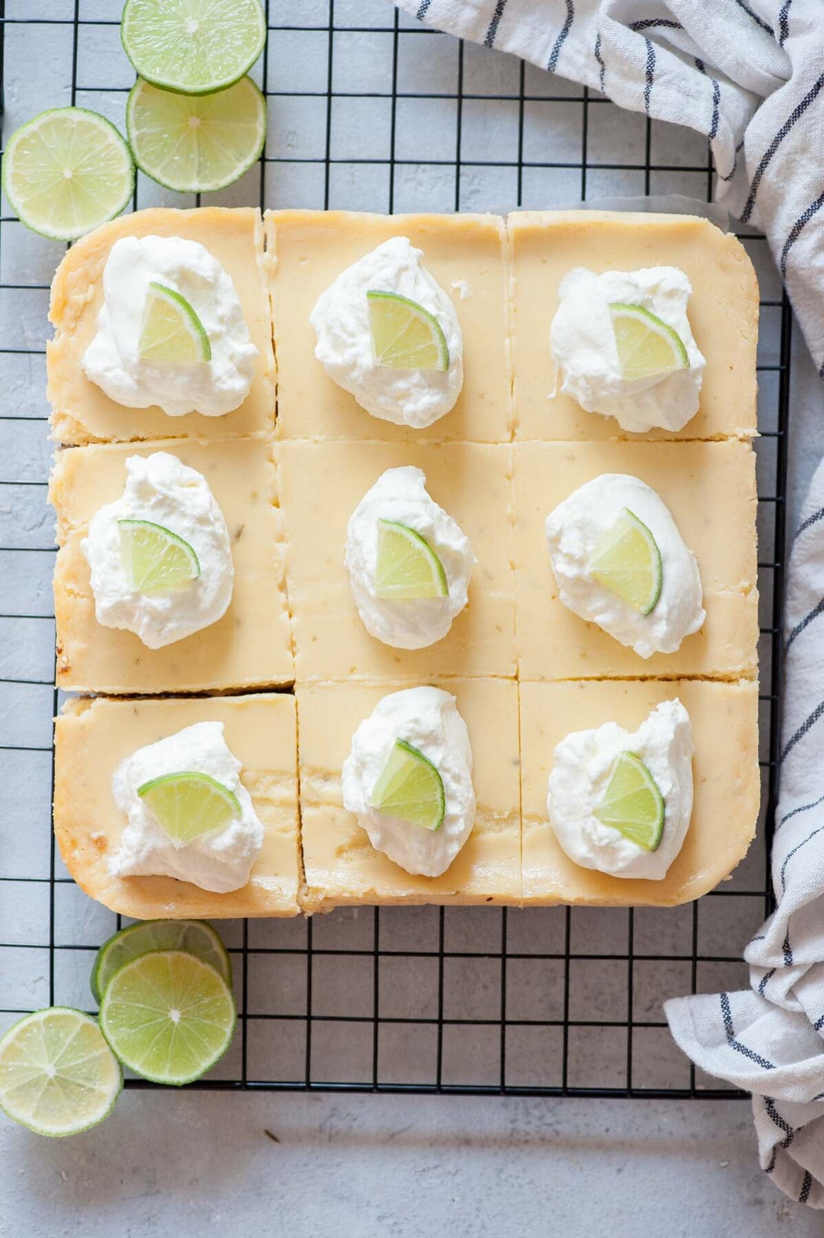 Key lime pie bars topped with whipped cream and lime slices on a black cooking rack.