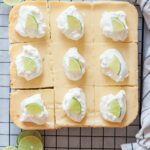 Key lime pie bars topped with whipped cream and lime slices on a cooling rack.