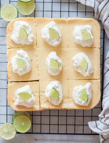 Key lime pie bars topped with whipped cream and lime slices on a cooling rack.