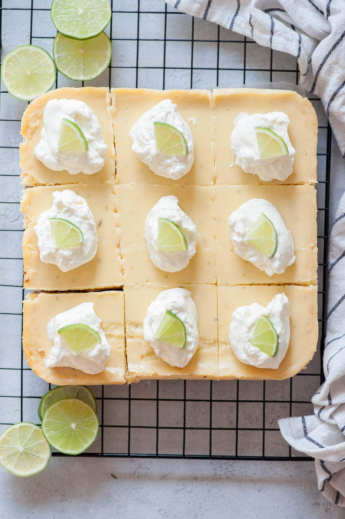 Key lime pie bars topped with whipped cream and lime slices on a cooling rack.