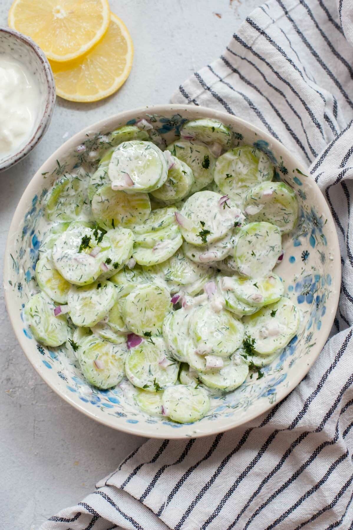 Mizeria (Polish cucumber salad) in a white bowl. Lemon slices and kitchen cloth in the background.