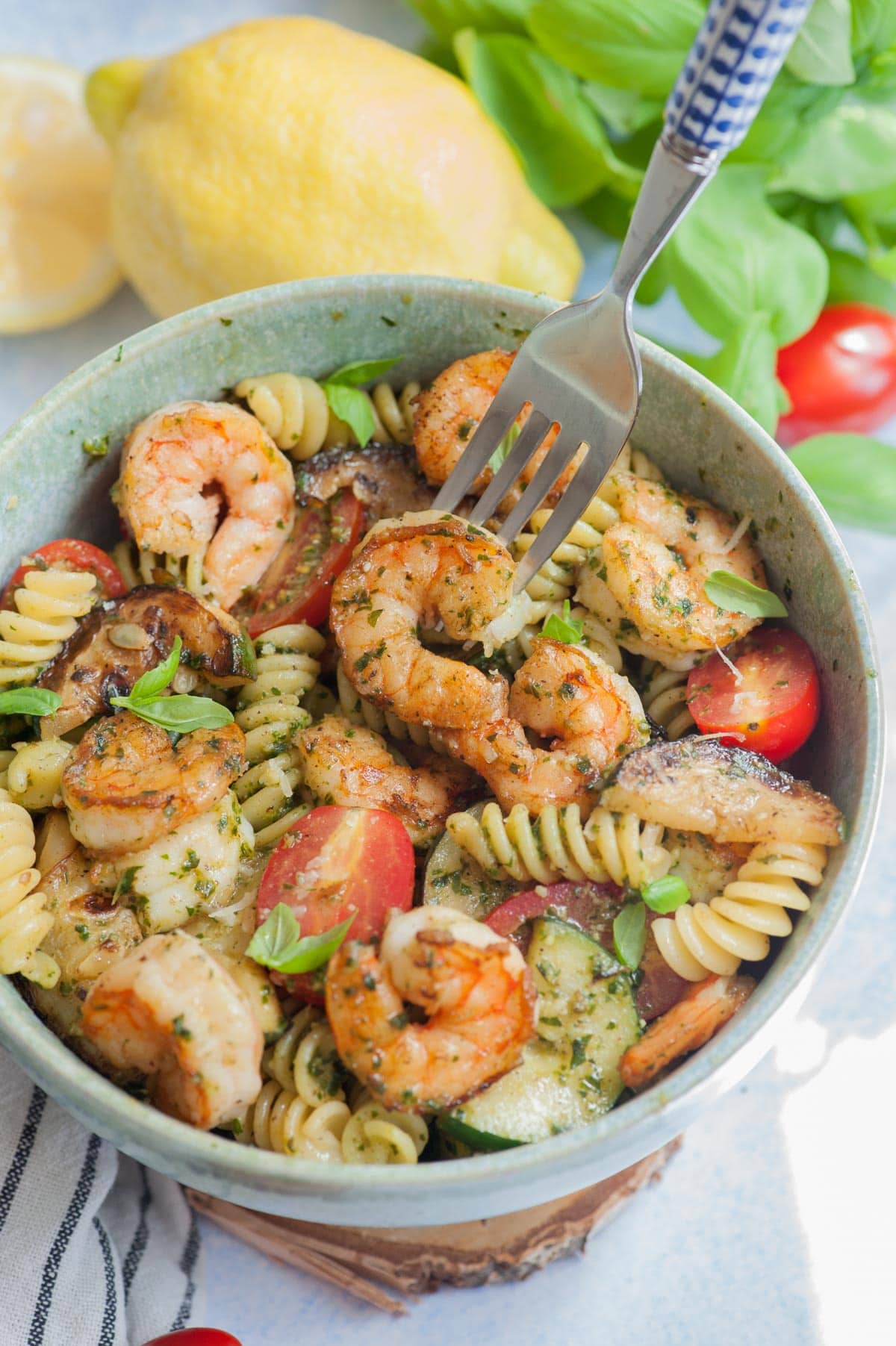 Shrimp pesto pasta in a green bowl. Basil leaves, lemons and tomatoes in the background.