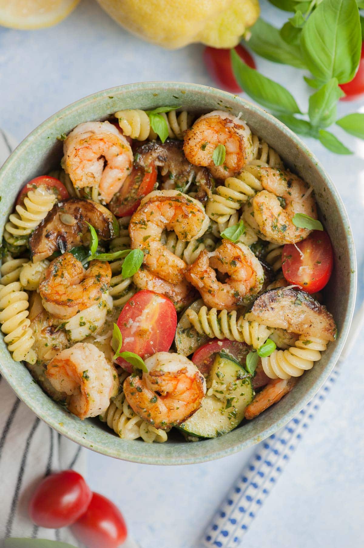 Shrimp pesto pasta in a green bowl. Basil leaves, lemons and tomatoes in the background.