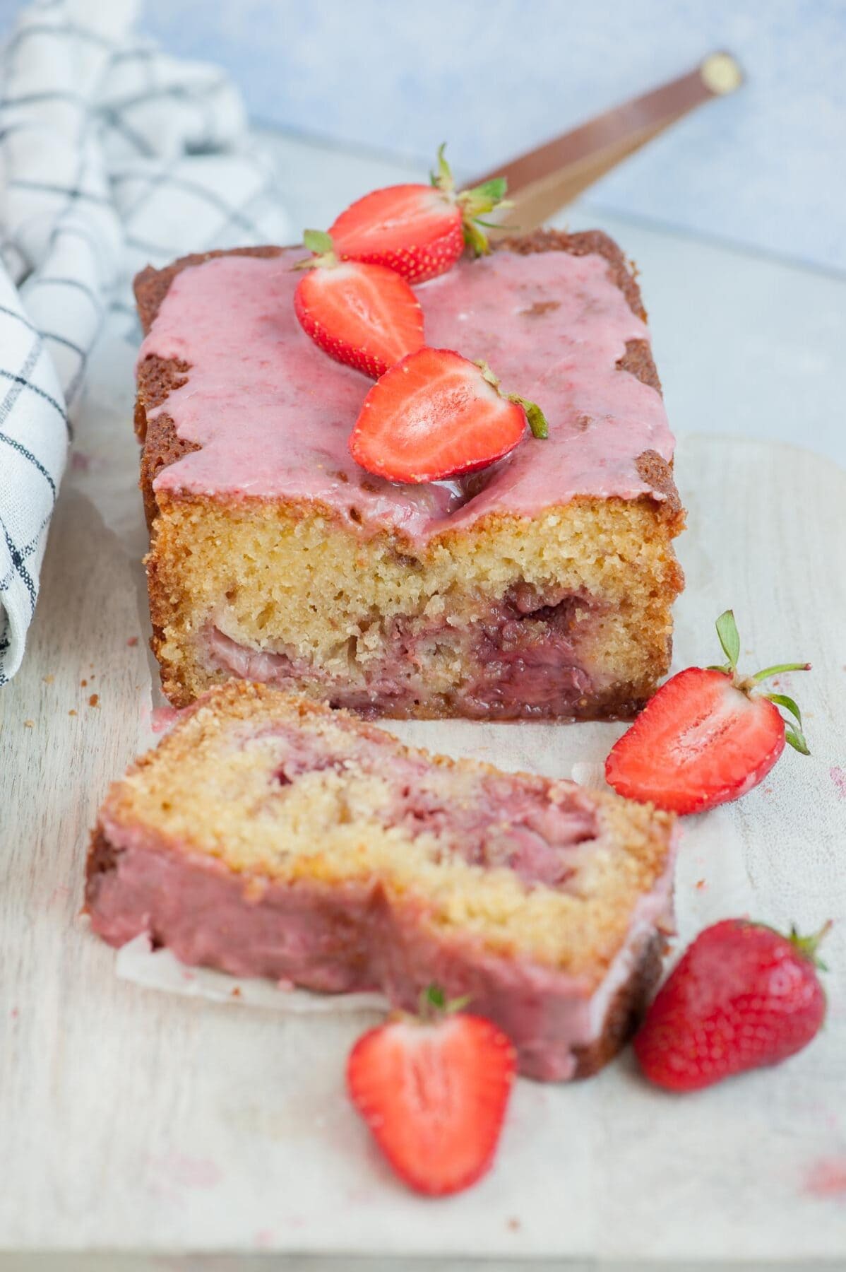 Strawberry bread with a slice cut off on a beige wooden board topped with strawberry icing and strawberries.
