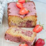 Strawberry bread with a slice cut off on a beige wooden board topped with strawberries.