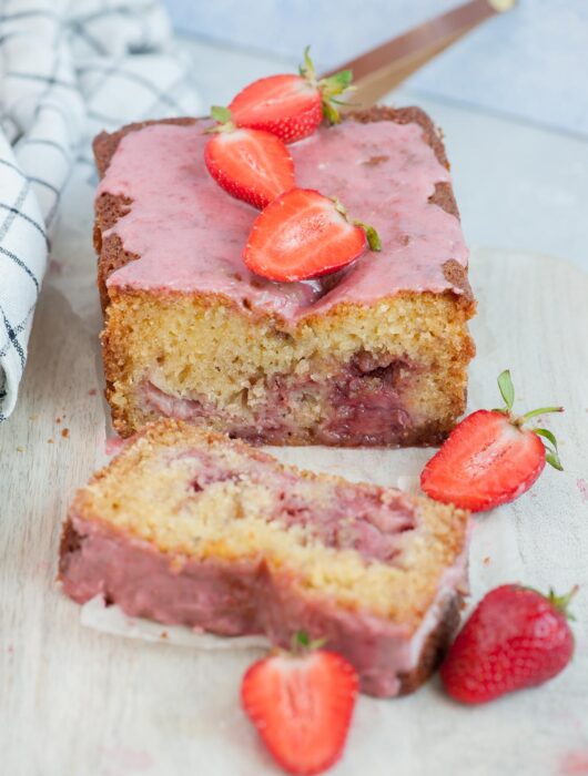 Strawberry bread with a slice cut off on a beige wooden board topped with strawberries.