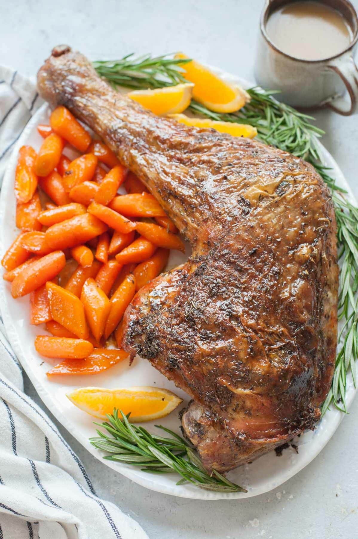 Whole baked turkey leg on a white plate with glazed carrots, rosemary springs and orange wedges.