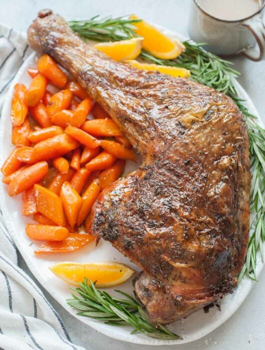 Whole roasted turkey leg on a white plate served with glazed carrots and potatoes.