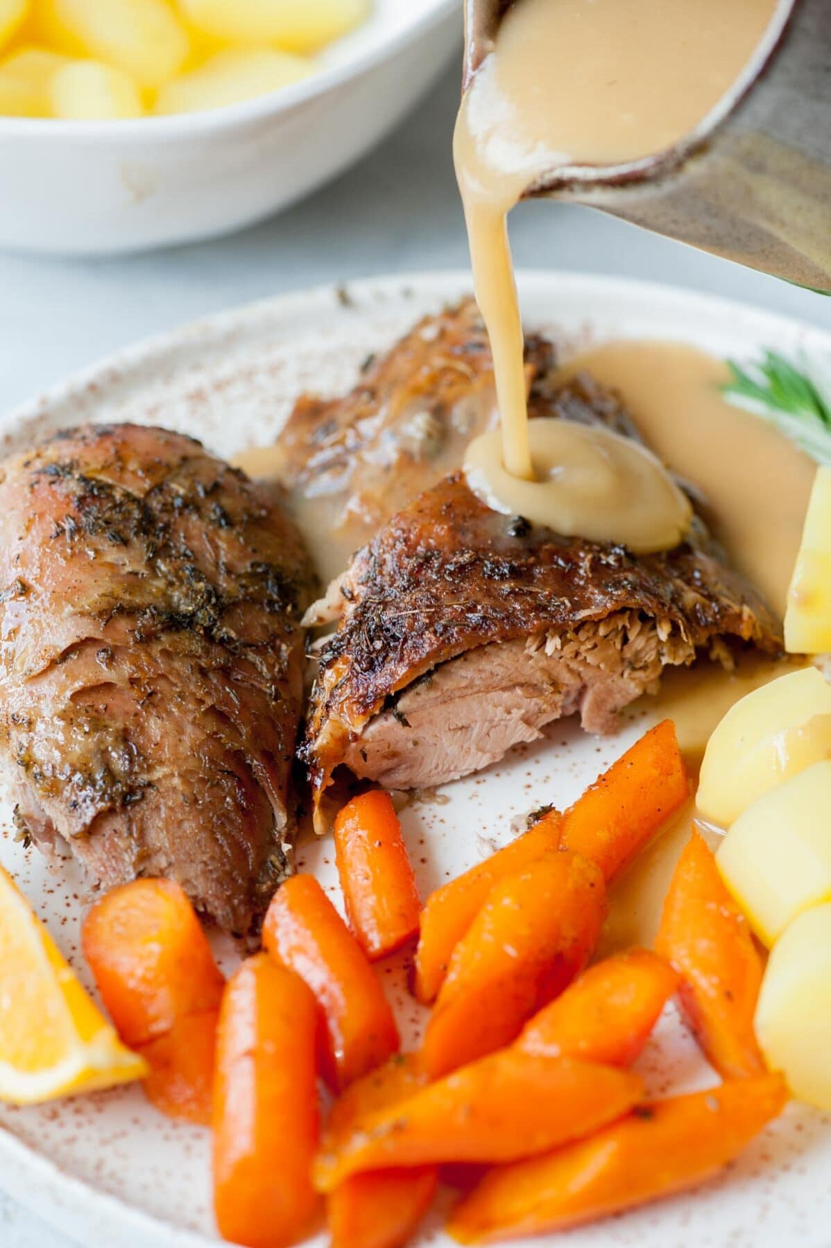 Gravy is being poured over baked turkey leg on a white plate with glazed carrots and potatoes.