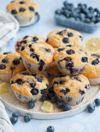 Blueberry muffins on a white plate. Blueberries and lemon slices scattered around.