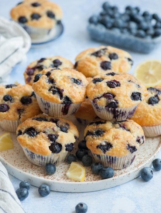 Blueberry muffins on a white plate. Blueberries and lemon slices scattered around.