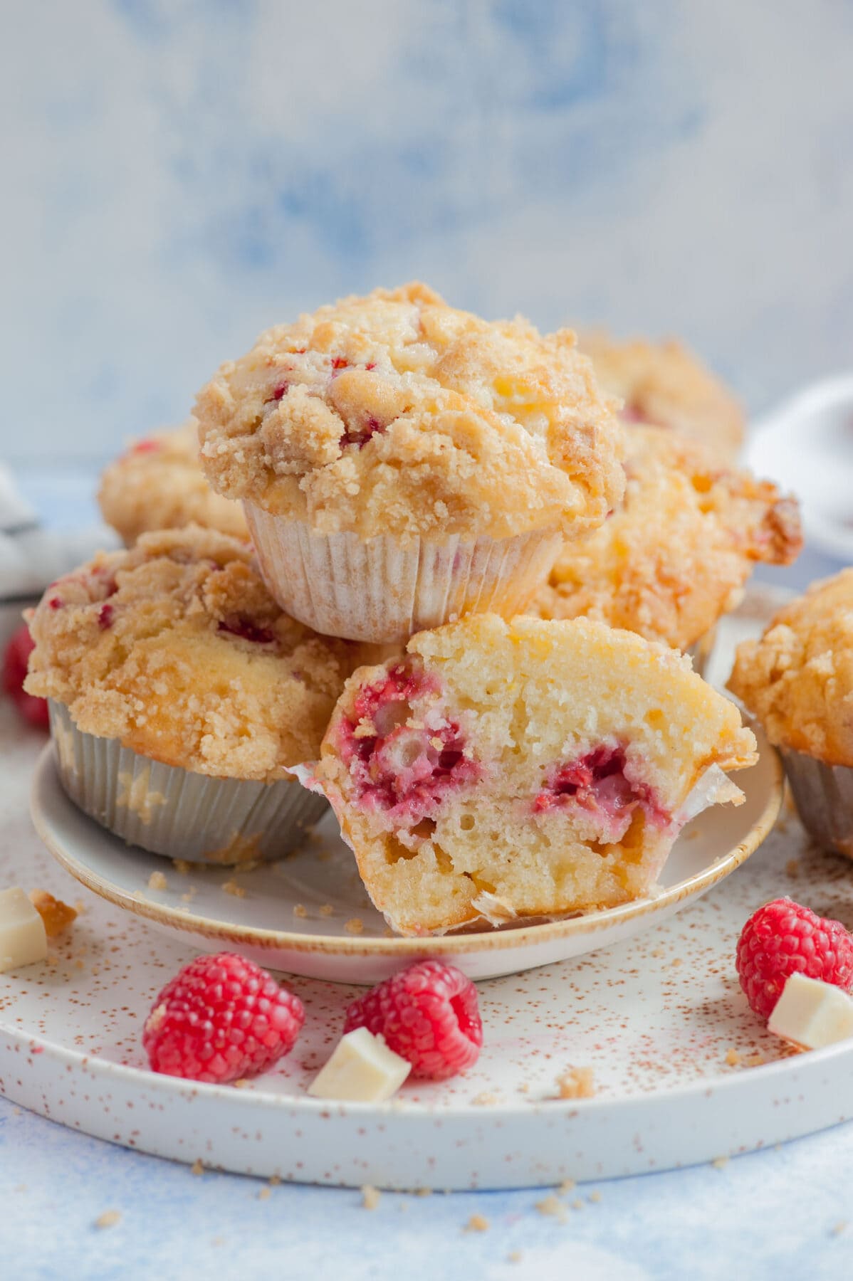 Raspberry muffins on a white plate. One muffin cut in half. Raspberries and white chocolate chunks scattered around.