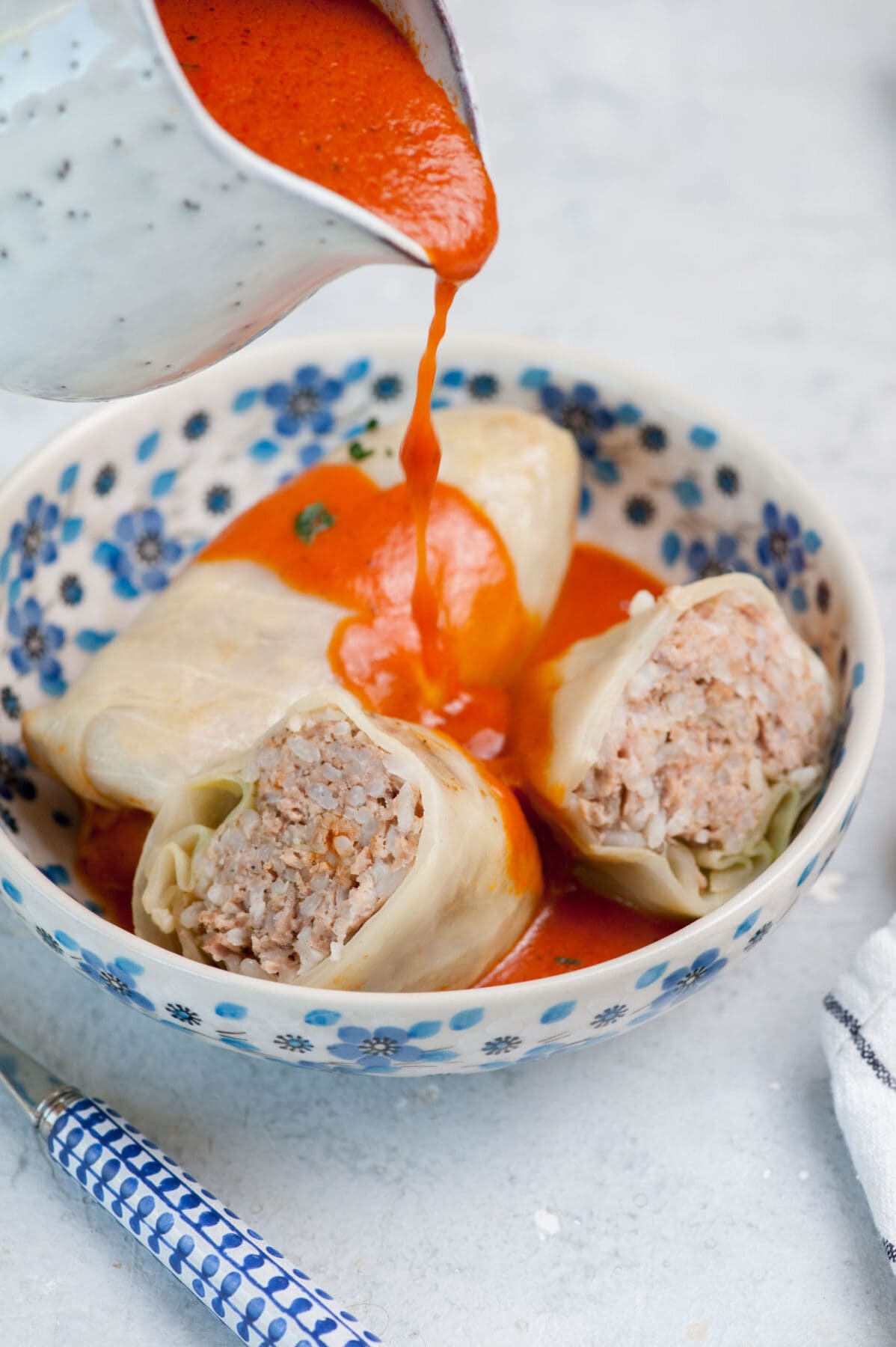 Tomato sauce is being poured over stuffed cabbage rolls in a white-blue bowl.