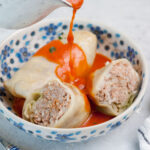 Tomato sauce is being poured over stuffed cabbage rolls in a white-blue bowl.