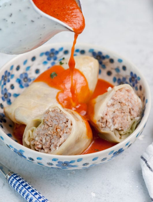 Tomato sauce is being poured over stuffed cabbage rolls in a white-blue bowl.