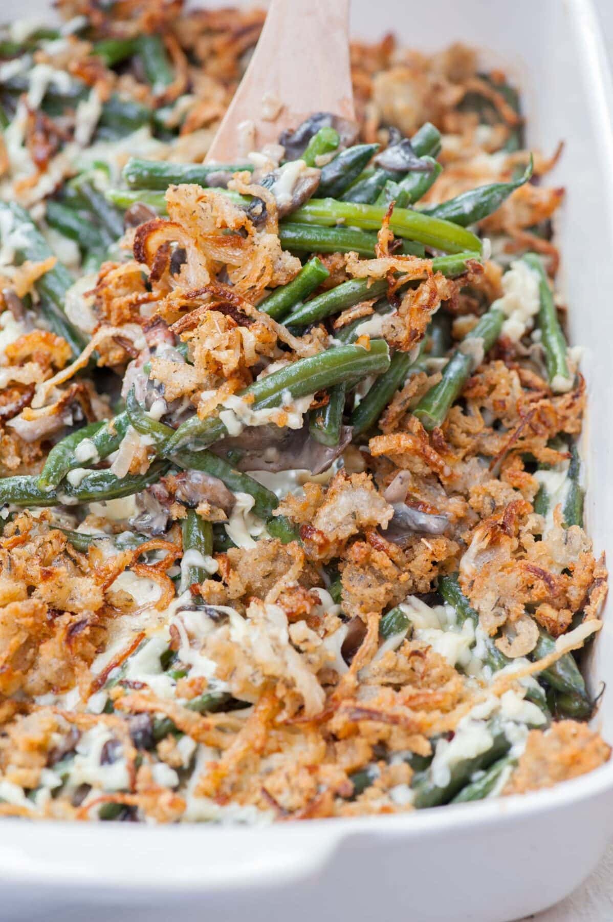 Green bean casserole in a white baking dish is being loaded on a wooden spatula.