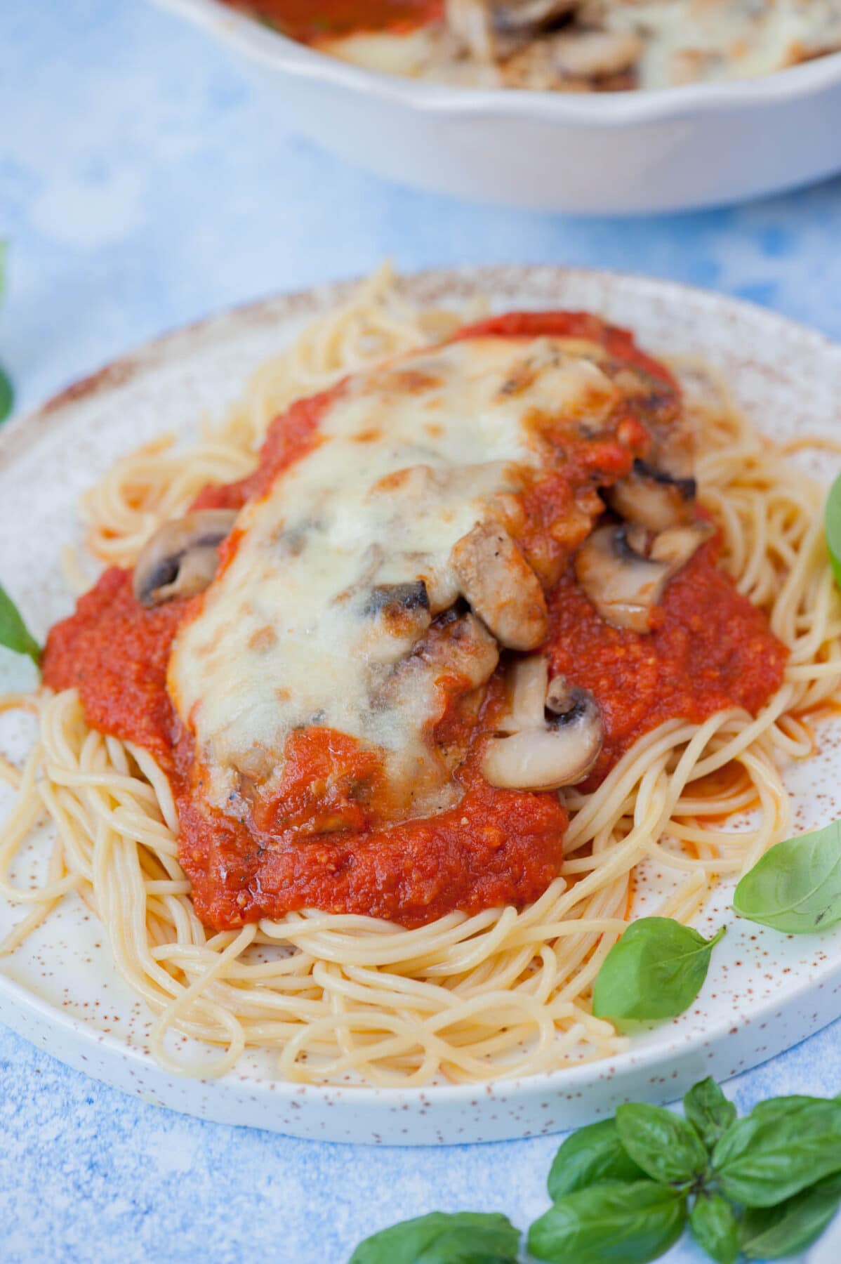 Mozzarella chicken with mushrooms and marinara sauce on top of spaghetti on a white plate.