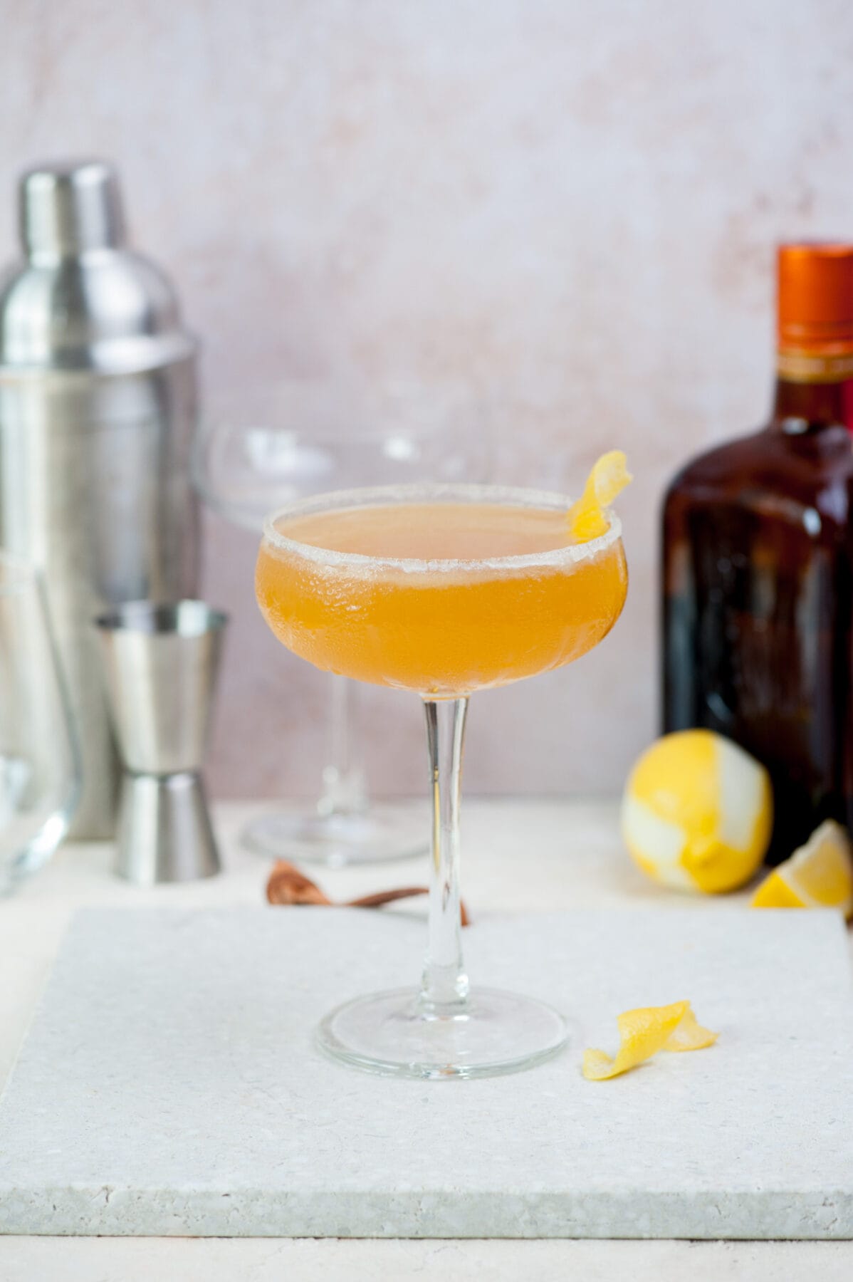 A coupe glass with sidecar cocktail on a grey stone board. A shaker and liqueur bottle in the background.