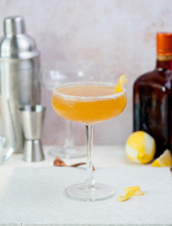 A coupe glass with sidecar cocktail on a grey stone board. A shaker and liqueur bottle in the background.