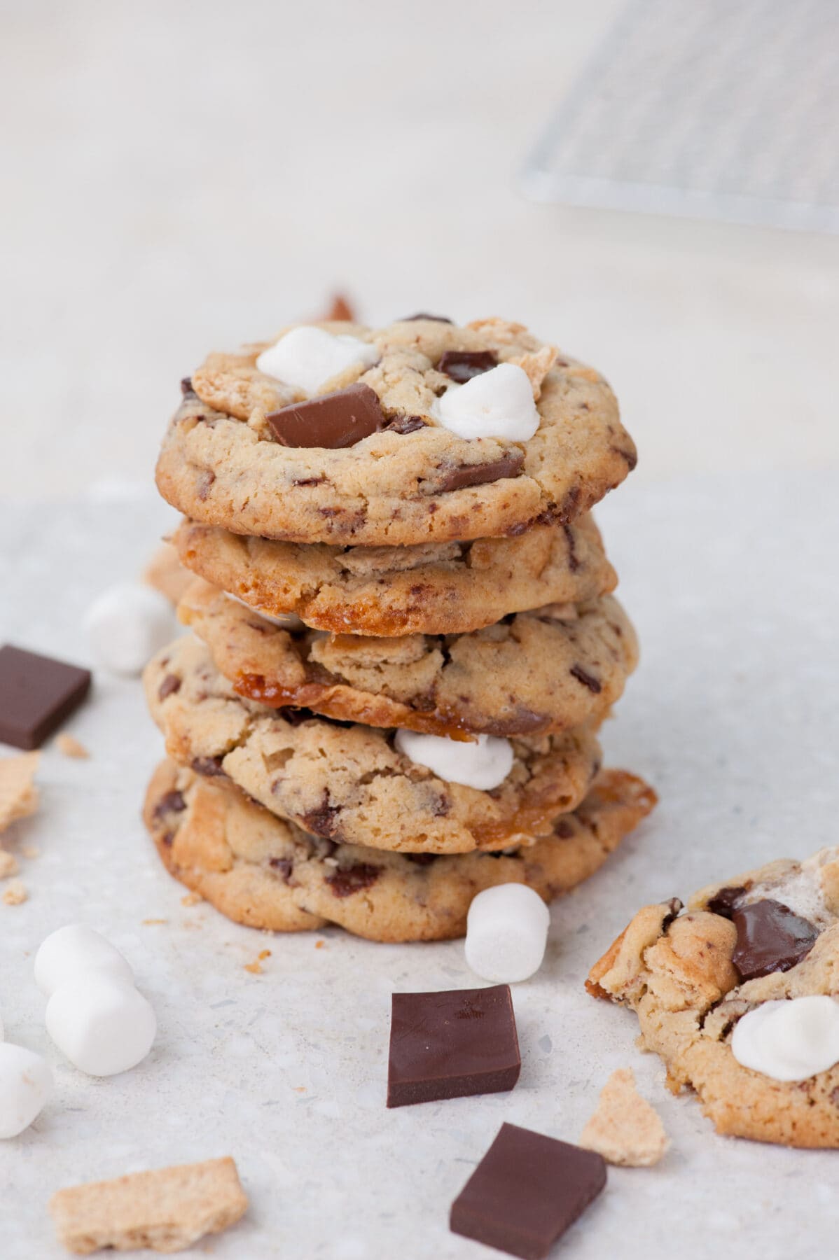 A stack of s'mores cookies on a grey background. Marshmallows and chocolate scattered around.