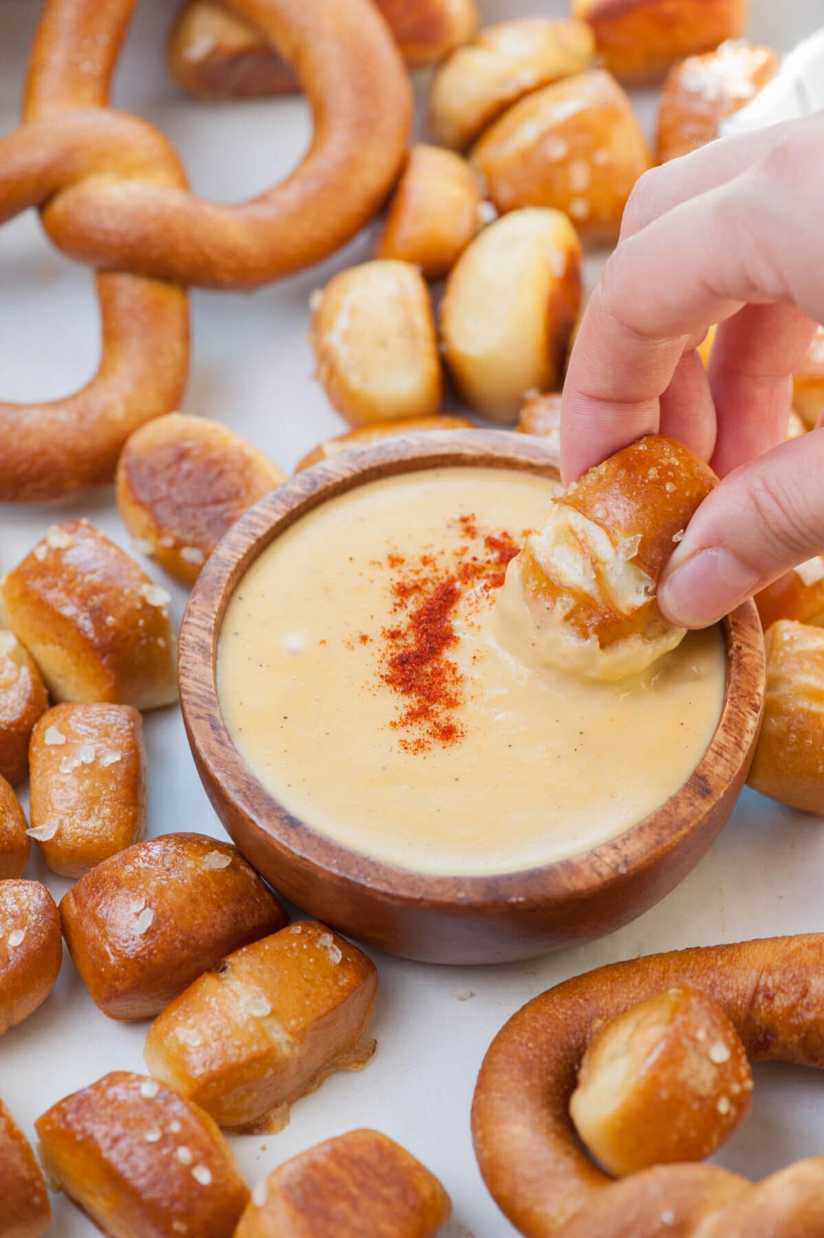 Soft pretzel bite is being dipped in a cheese sauce in a brown bowl.. Pretzel bites scattered around.