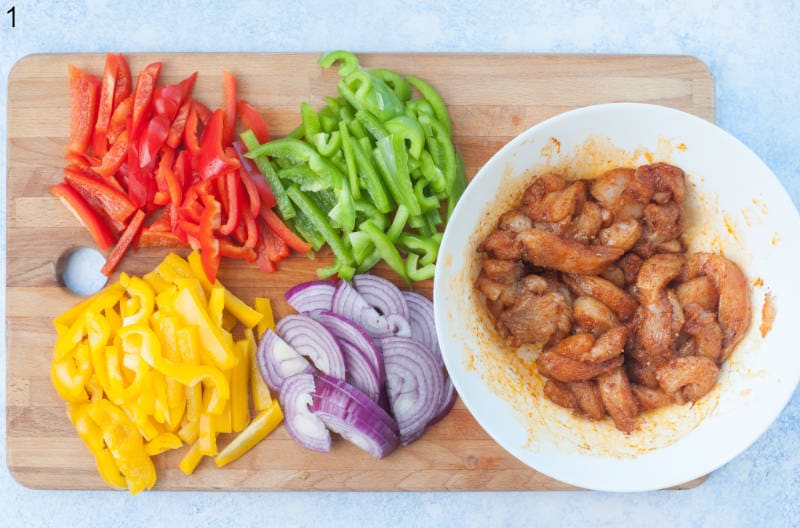 Chopped bell peppers and red onion on a chopping board. Strips of chicken with Fajita seasoning in a white bowl.
