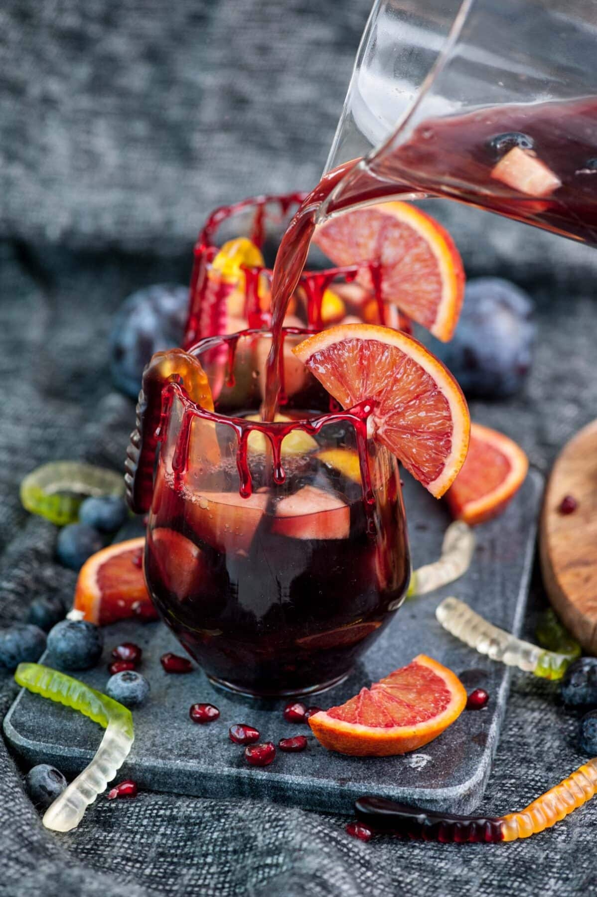 Sangria is being poured into two glasses garnished with blood orange slices and fake blood drips.