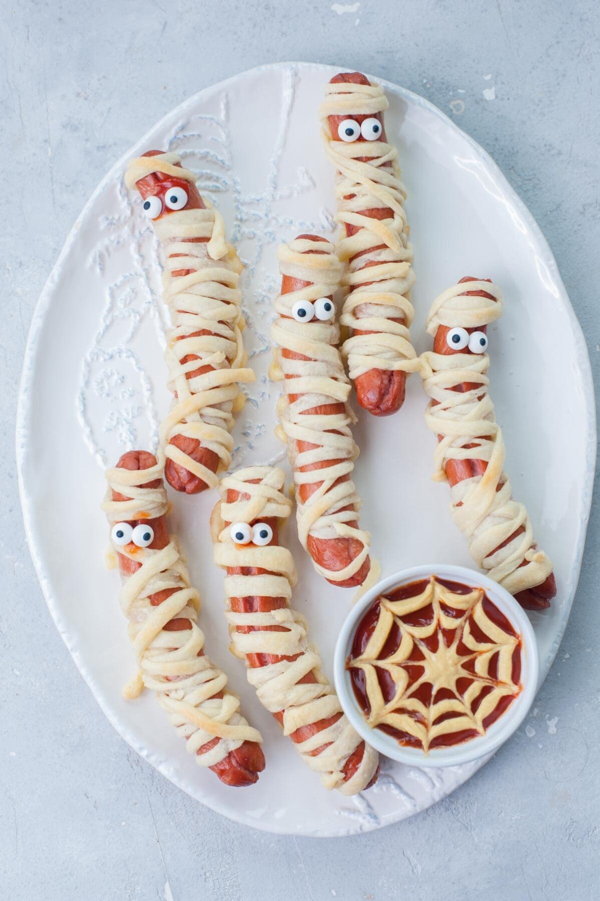 Mummy hot dogs on a white plate with ketchup mustard dip in a small bowl.