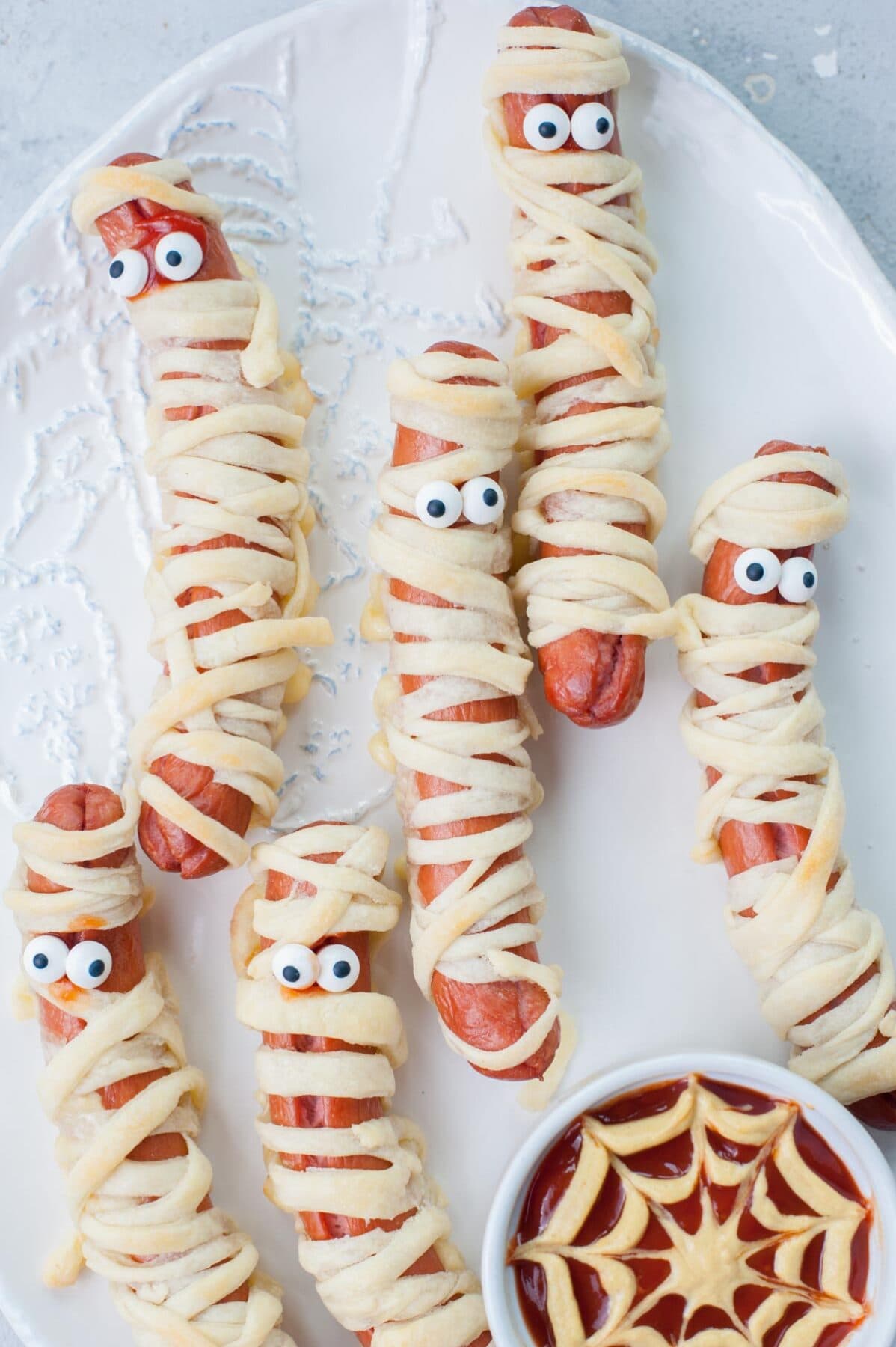 Mummy hot dogs on a white plate with ketchup mustard dip in a small bowl.