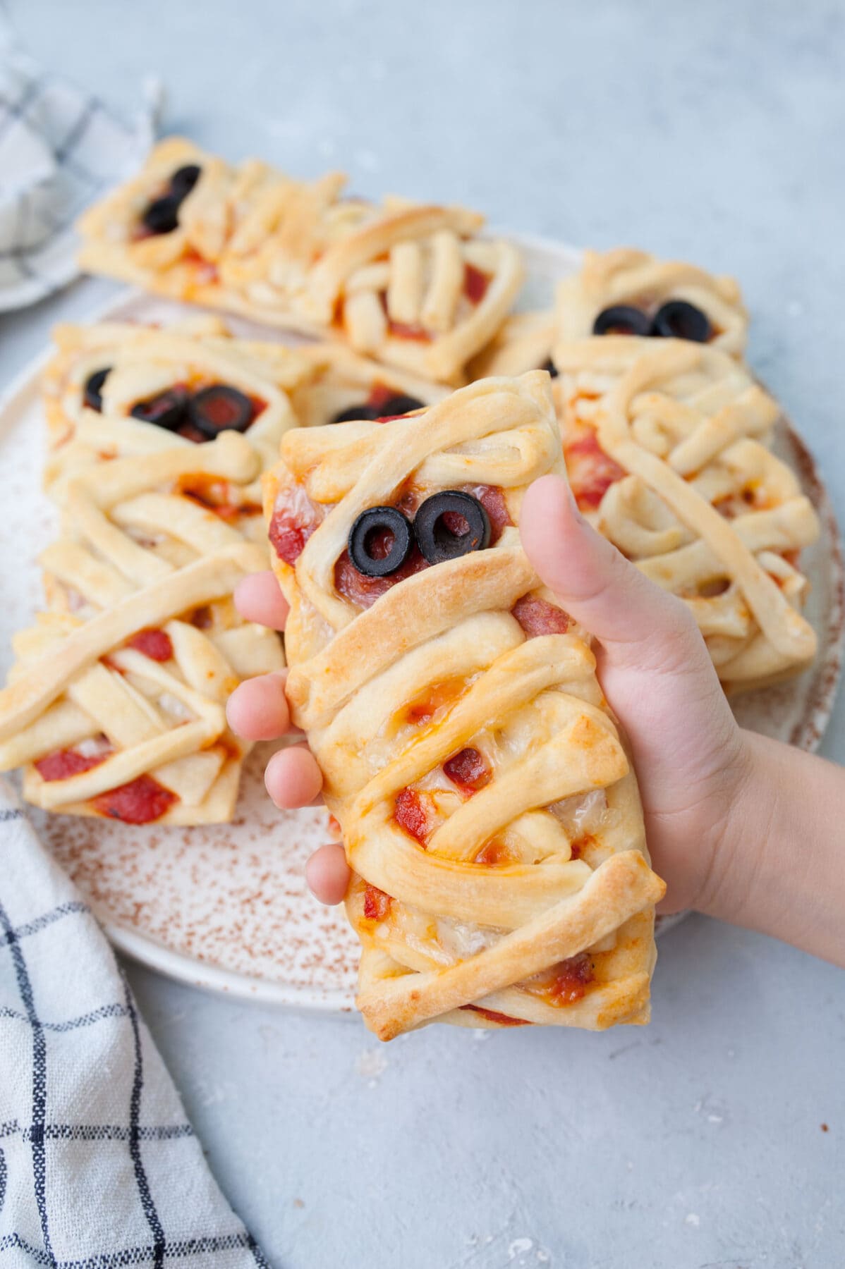 A pizza mummy held in a child's hand. More pizza mummies on a plate in the background.