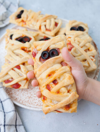 Pizza mummy held in a child's hand. More pizza mummies on the plate in the background.